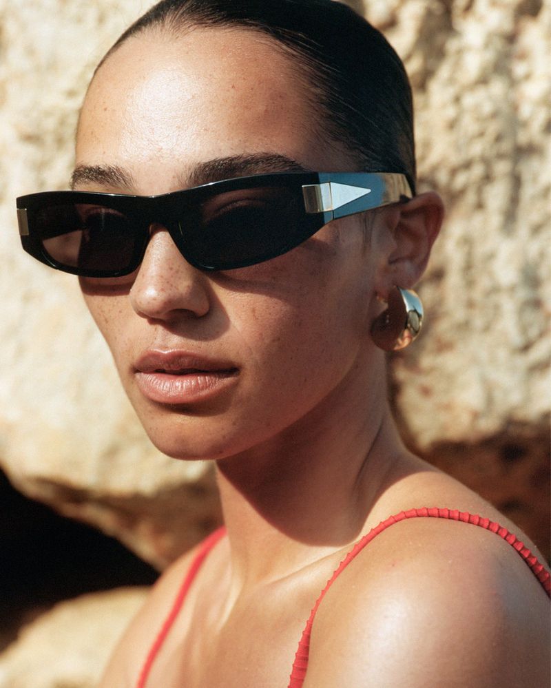 Close-up of a woman wearing sunglasses and gold hoop earrings, standing in front of a light-brown rock wall in natural light.