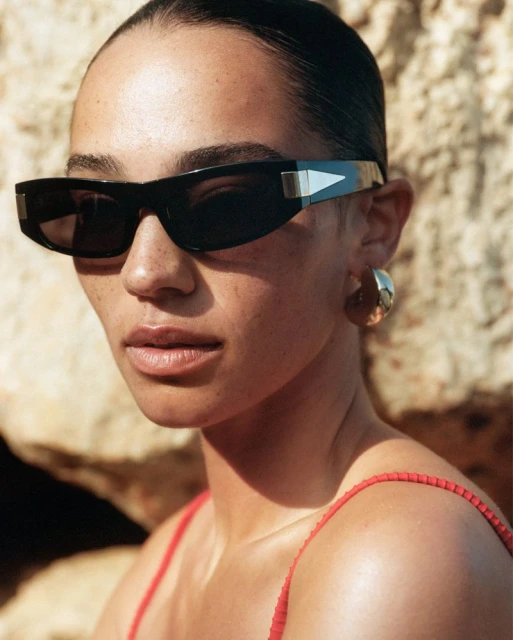Close-up of a woman wearing sunglasses and gold hoop earrings, standing in front of a light-brown rock wall in natural light.