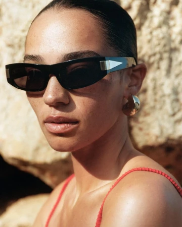 Close-up of a woman wearing sunglasses and gold hoop earrings, standing in front of a light-brown rock wall in natural light.