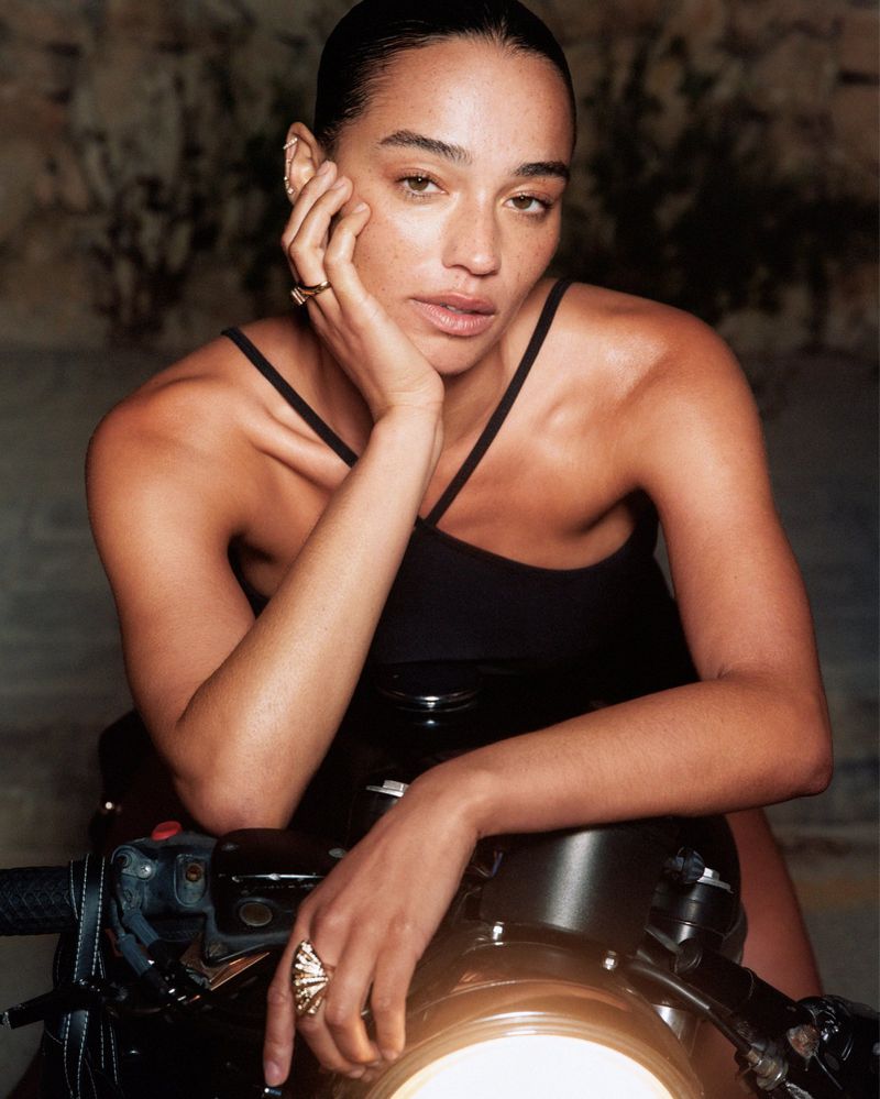 Adult woman with fair skin poses with hand on her face while sitting on a black motorcycle in an indoor location with soft lighting.