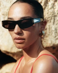 Close-up portrait of a woman wearing black sunglasses and a red textured top against a rocky textured background.