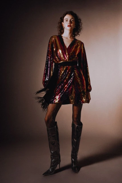 A stylish woman with curly hair poses in a studio wearing a sequin dress, patterned tights, and black high-heeled boots, fashion shoot.