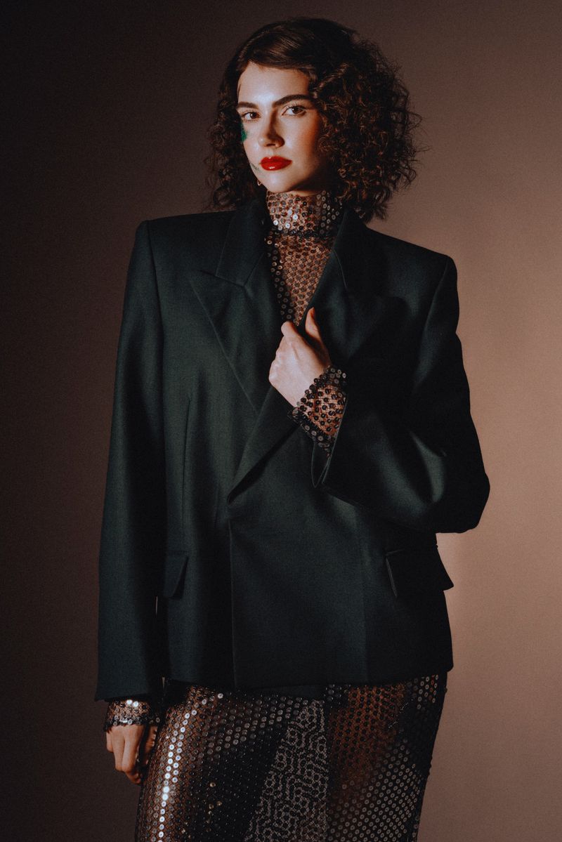 Elegant woman in a black blazer and sequined skirt poses in a studio with soft lighting, showcasing fashion and style.