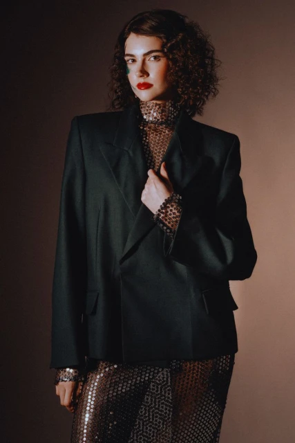 Elegant woman in a black blazer and sequined skirt poses in a studio with soft lighting, showcasing fashion and style.