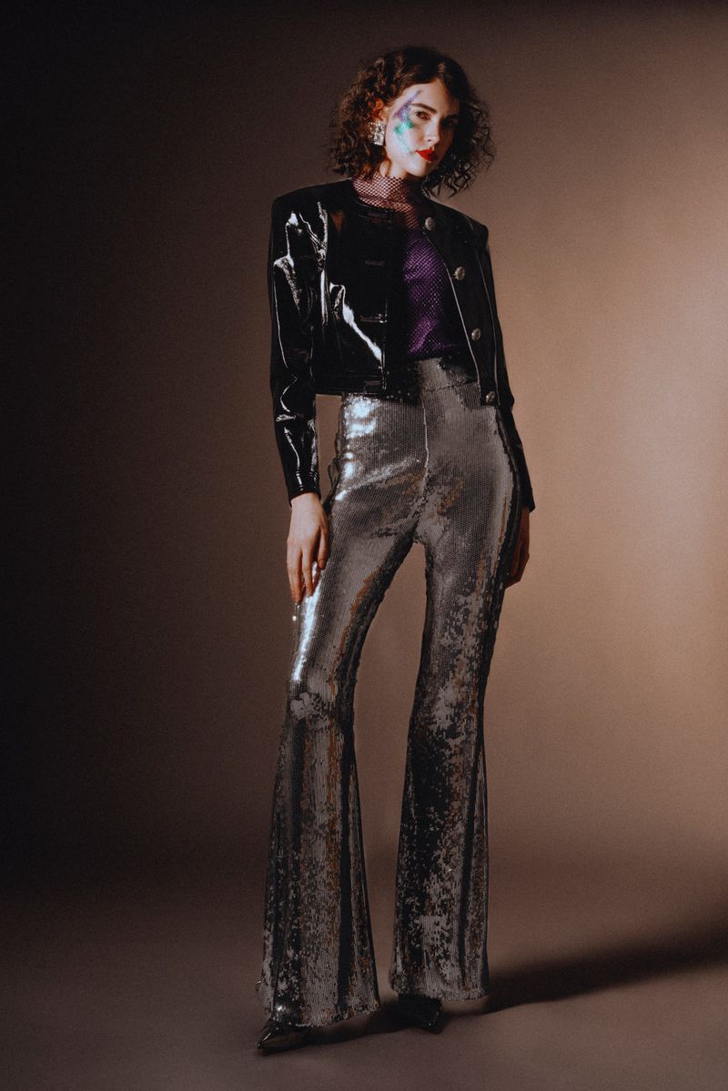 A woman in a shiny black jacket and shimmering bell-bottoms poses against a neutral backdrop, showcasing a striking fashion style.