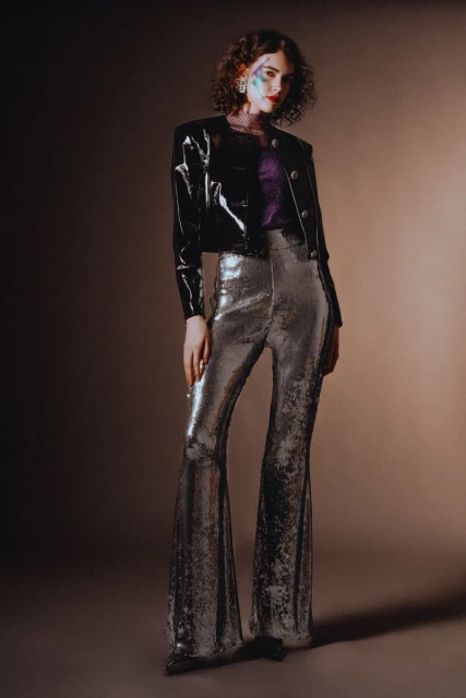 A woman in a shiny black jacket and shimmering bell-bottoms poses against a neutral backdrop, showcasing a striking fashion style.