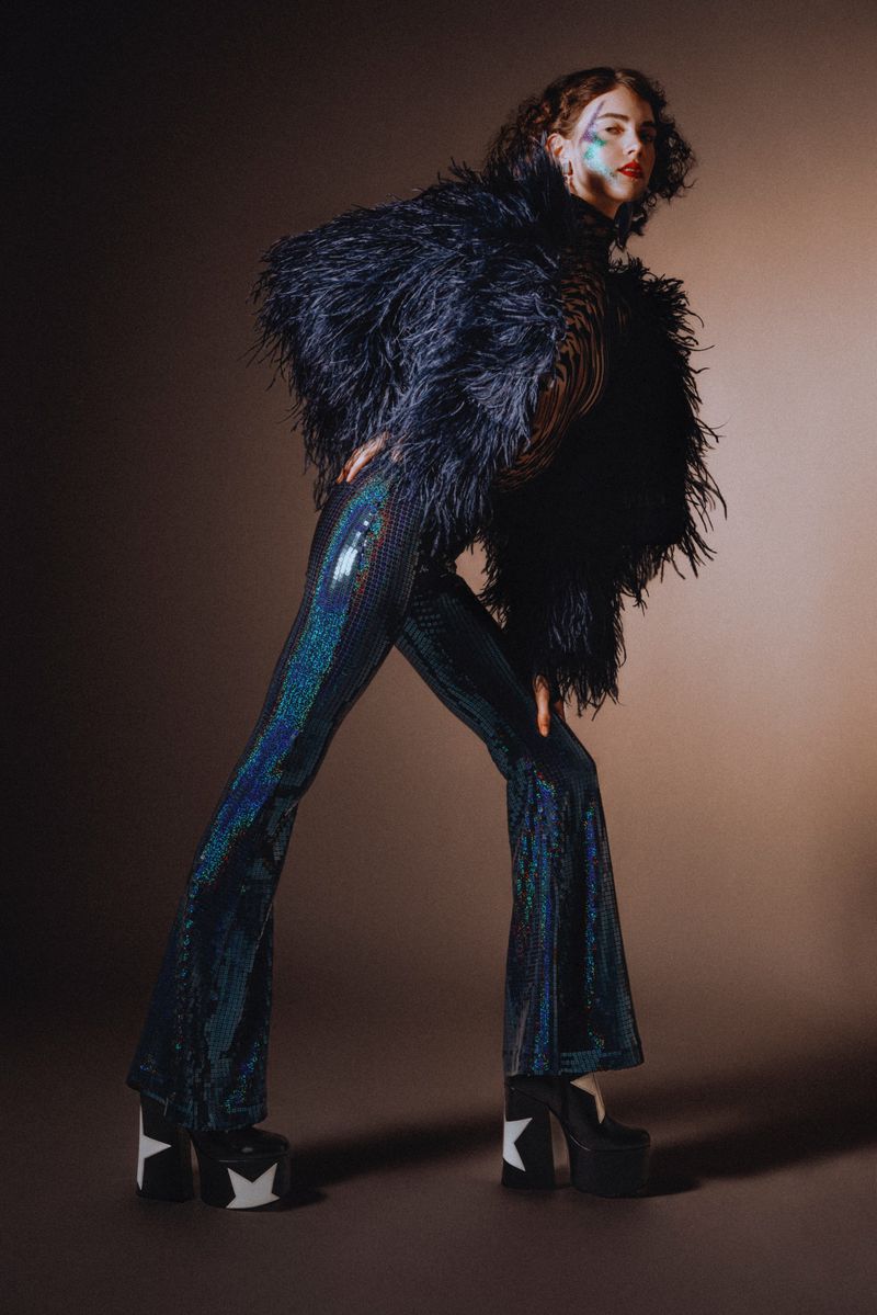 Woman in feathered jacket and iridescent pants poses against a graduated brown backdrop. Platform shoes with stars add to the look.