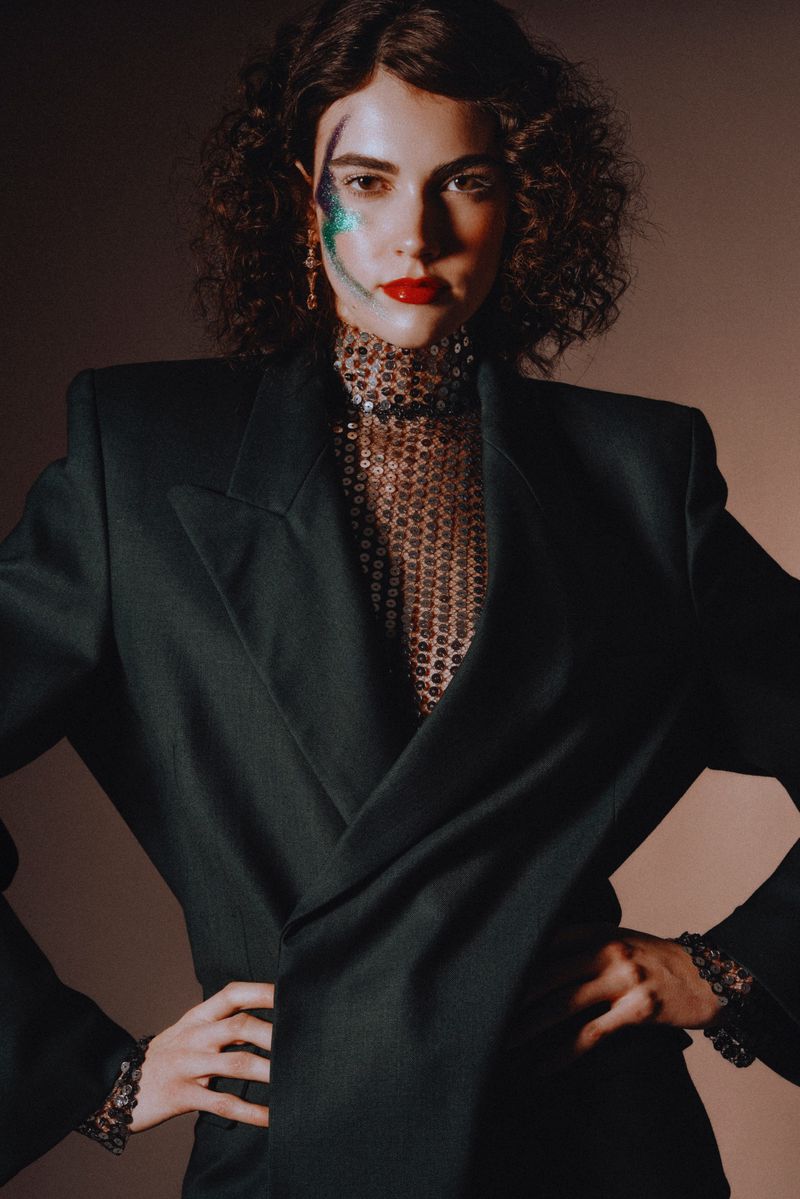 Stylish woman in a blazer and sequined top strikes a pose with hands on hips, showcasing bold makeup against a neutral backdrop.