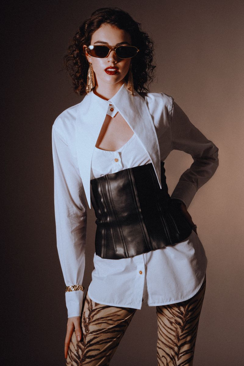 Fashionable woman poses in a white button-down shirt with a black corset, tiger-print leggings, and sunglasses, with a neutral background.