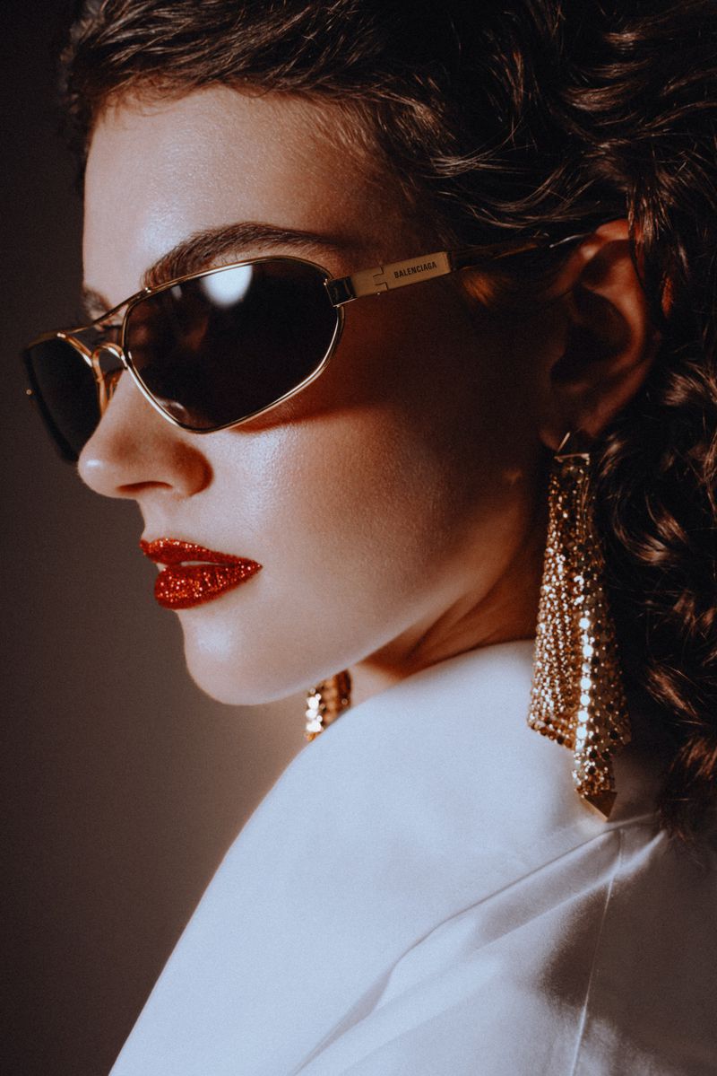 Stylish woman poses in close-up wearing sunglasses and large chandelier earrings with bright red glitter lipstick and a white jacket.