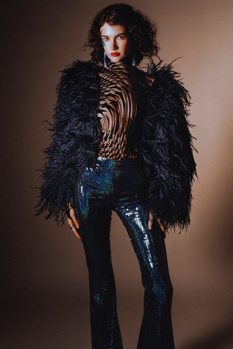 A woman poses in a fashionable feather jacket and sequined pants against a neutral backdrop, showcasing a contemporary style.