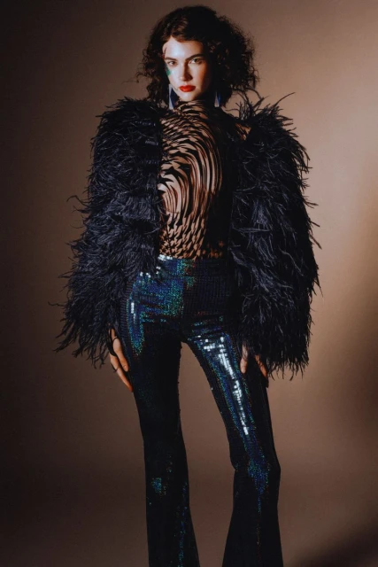 A woman poses in a fashionable feather jacket and sequined pants against a neutral backdrop, showcasing a contemporary style.