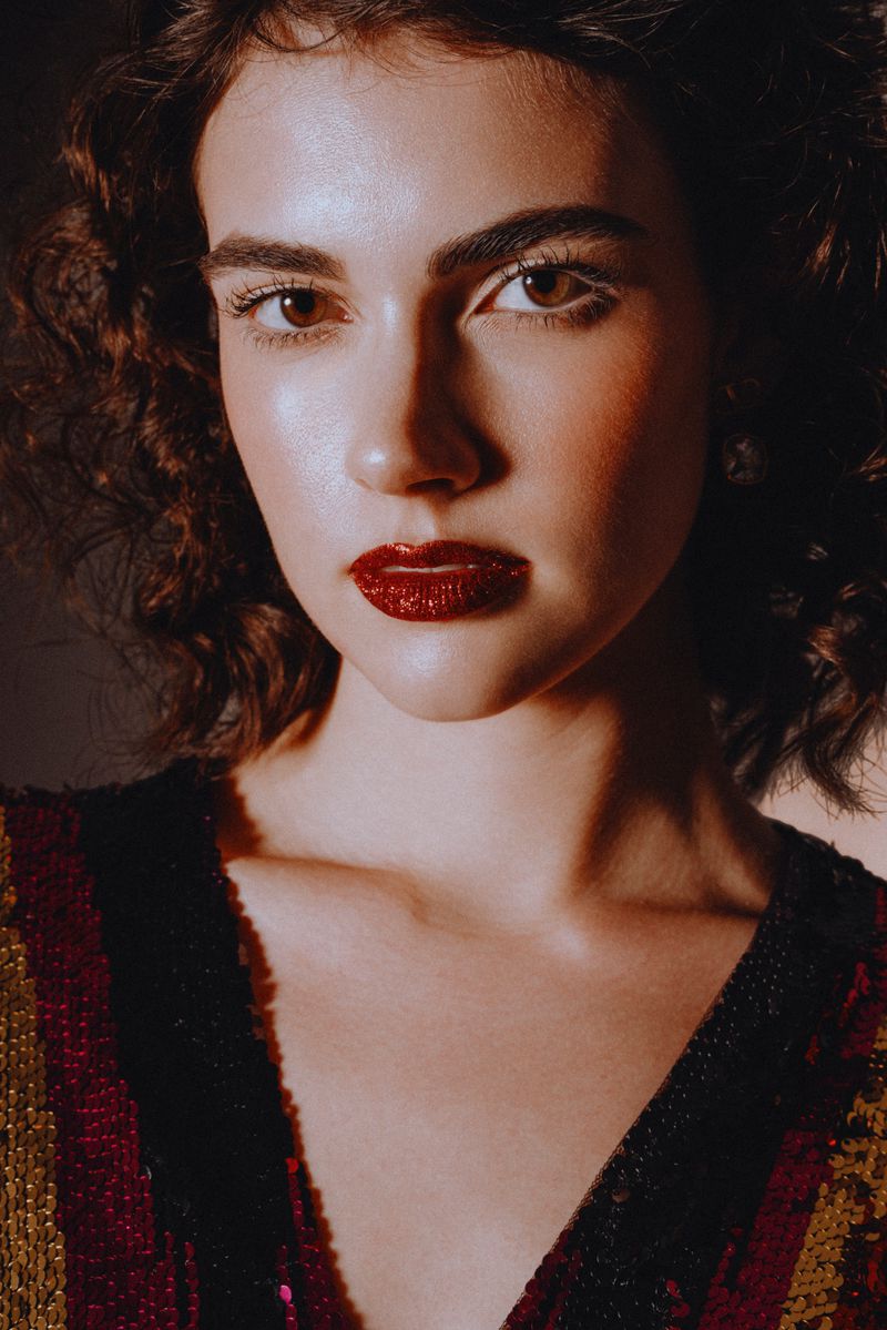 Close-up portrait of a young adult woman with dark curly hair, wearing a red glitter lipstick and a sequined top in a dimly lit indoor setting.