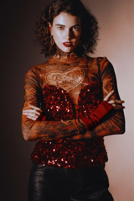 Portrait of an adult woman in a long-sleeved graphic top with red sequin detail, black leather pants, and curly hair in a studio setting.