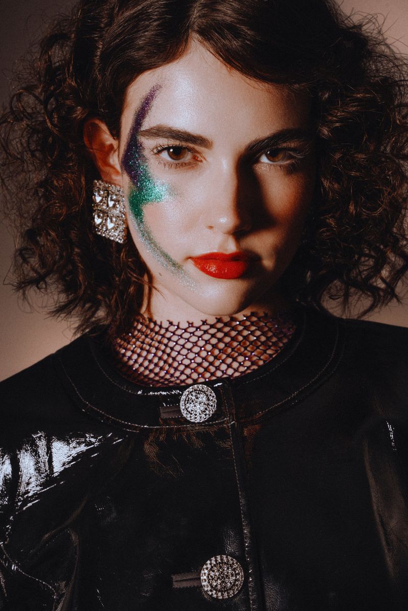 Close-up studio portrait of a young woman with dark curly hair, wearing a glossy black jacket with decorative buttons and bold eye makeup.