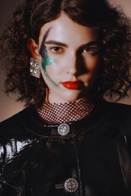 Close-up studio portrait of a young woman with dark curly hair, wearing a glossy black jacket with decorative buttons and bold eye makeup.