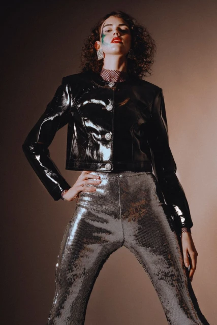 Fashionable adult female model posing in a shiny black jacket and silver sequin pants in a studio, illuminated by directed lighting.