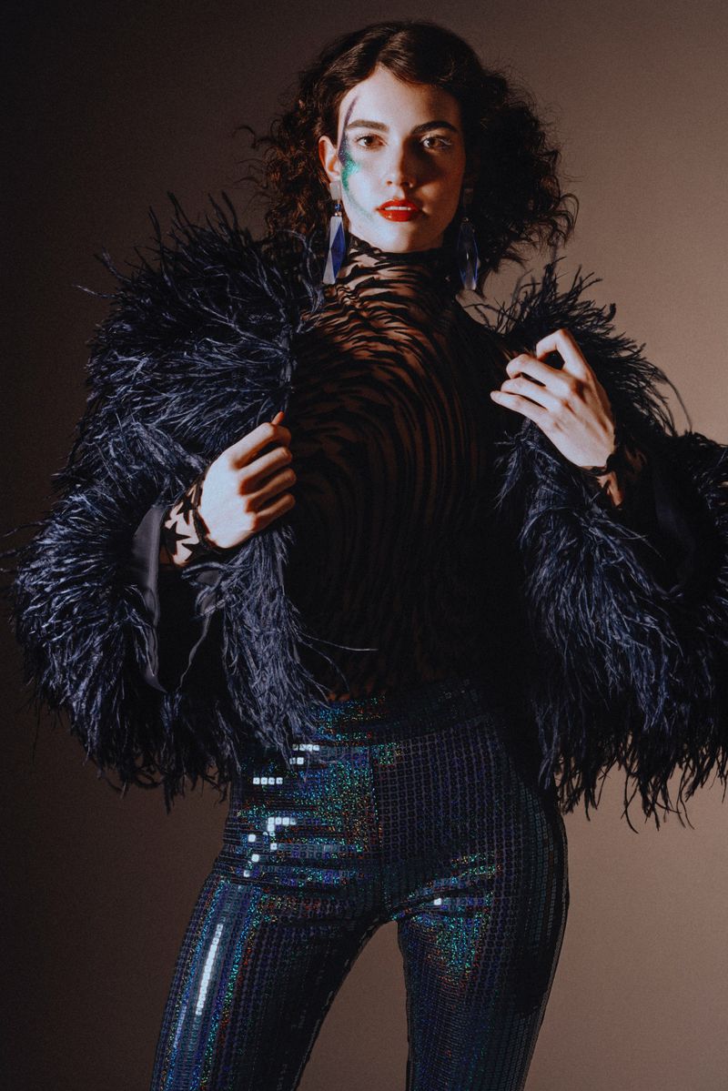 Elegant woman in feathered jacket and sequined pants posing in photo studio with face makeup and dark curly hair against brown backdrop.