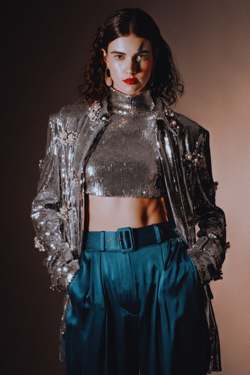 Brunette woman poses in a silver sequin top and jacket with teal satin pants against a tan backdrop, showcasing a retro-inspired fashion look.