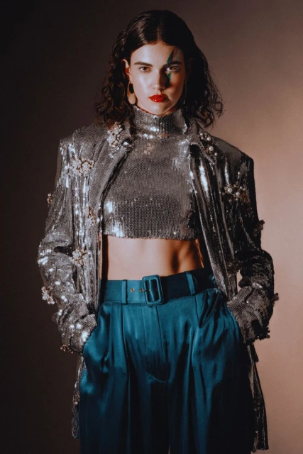 Brunette woman poses in a silver sequin top and jacket with teal satin pants against a tan backdrop, showcasing a retro-inspired fashion look.