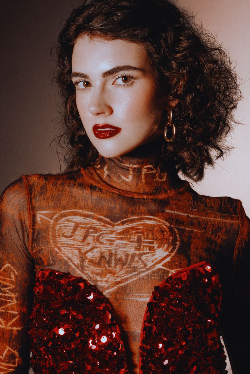 Portrait of a young woman with curly dark hair, wearing a sheer patterned top and a red sequined bodice, posing against a neutral background.