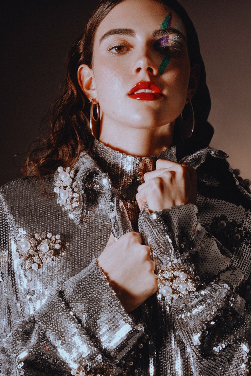 A woman poses in a sequined jacket, accented with rhinestones and pearls. She has artistic makeup and red lips against a dark background.