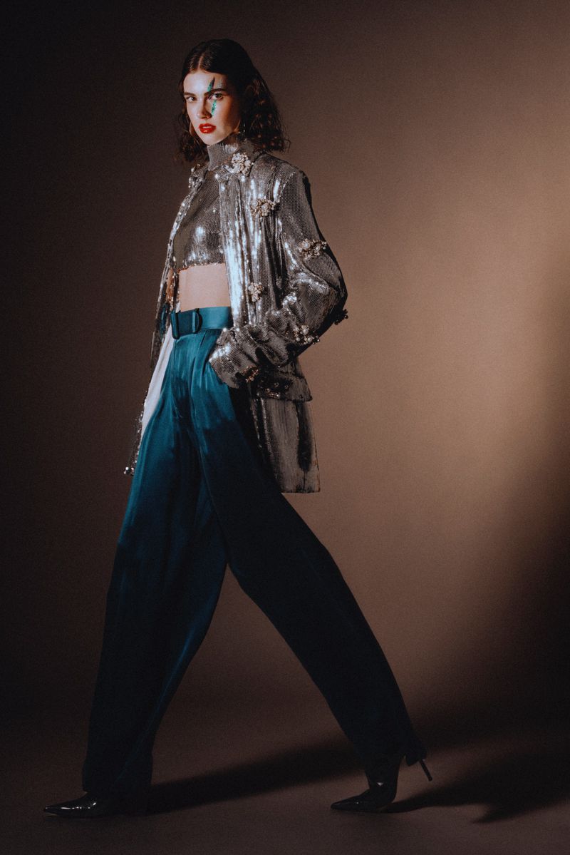 Woman in sequin top and teal pants strikes a pose with makeup in a fashion studio. The backdrop is a warm tan tone.