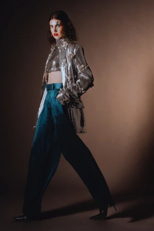 Woman in sequin top and teal pants strikes a pose with makeup in a fashion studio. The backdrop is a warm tan tone.