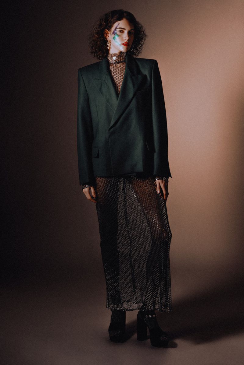 Model with dark curly hair and face paint wearing an oversized dark green blazer and sheer mesh skirt