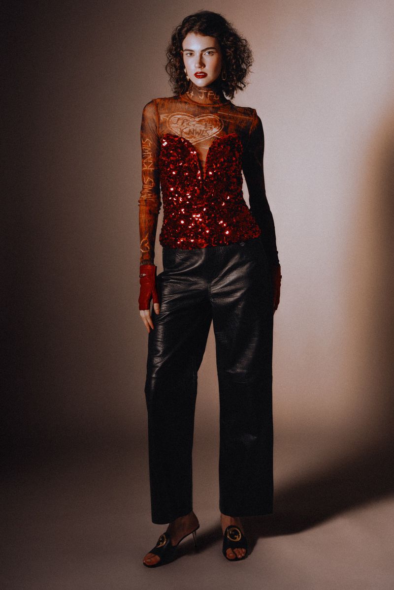 Woman poses in a sheer long-sleeved top with a red sequin bodice, paired with black leather pants and sandal heels in a studio shot.