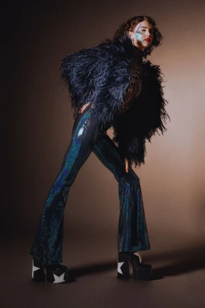Model in voluminous feather jacket and shiny patterned flare trousers posing against a brown studio backdrop