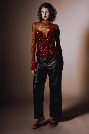 Model with dark curly hair wearing a sheer patterned top, sequined red bustier, and black leather trousers against a neutral background.