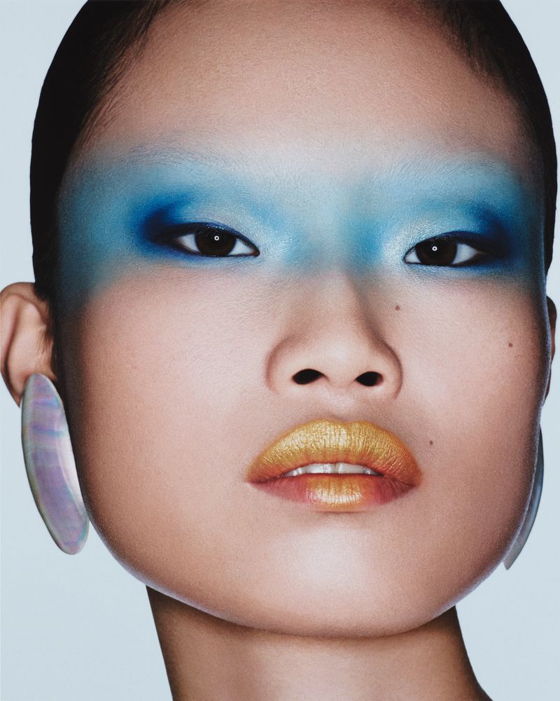 Close-up portrait of a woman with golden lips and blue eye makeup, wearing large oval iridescent earrings against a light blue background.