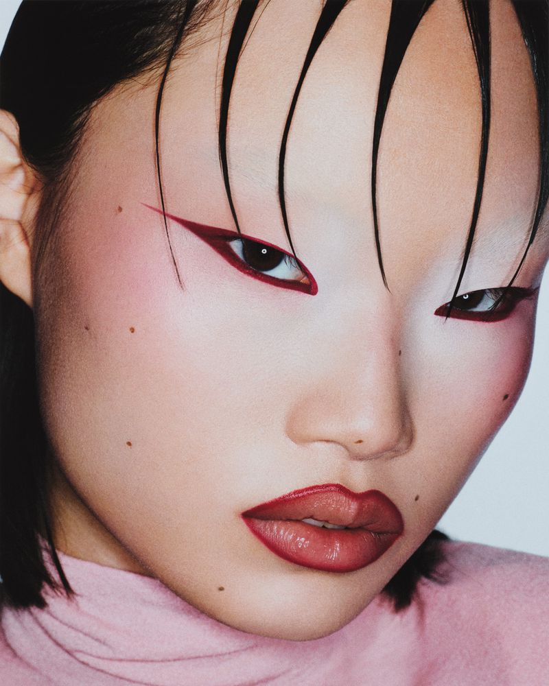 Close-up portrait of an Asian woman with wet, styled hair, distinctive red eyeliner and lipstick, fair skin, and wearing a pink top.