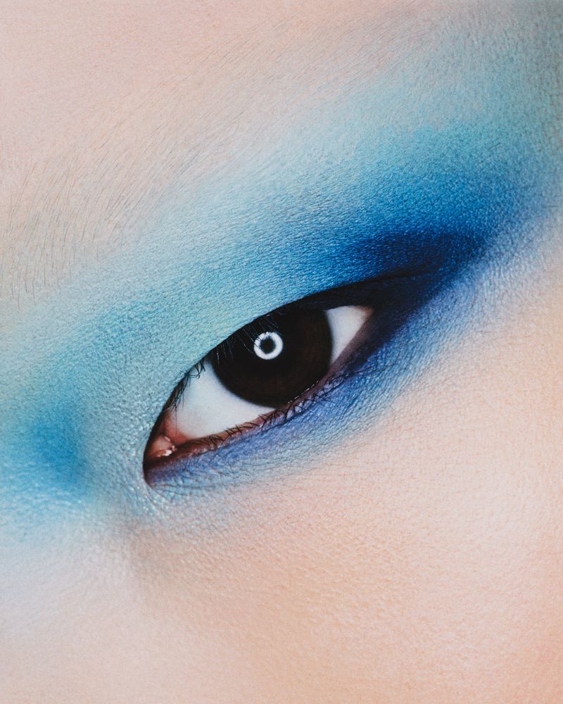 Close-up of a person's eye with blue eyeshadow. Focus on the dark pupil, surrounded by light skin tone.