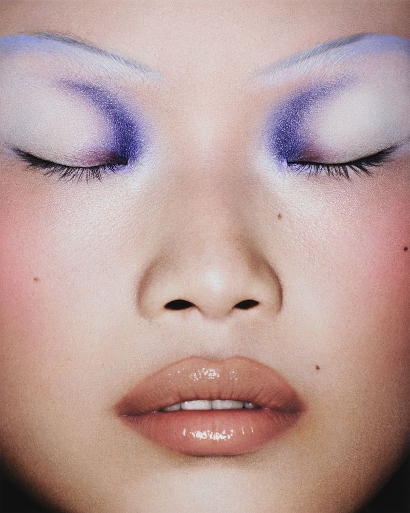 Close-up portrait of an Asian female face with closed eyes, glossy lips, and purple eyeshadow makeup for a beauty and fashion shot.