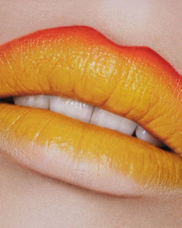 Extreme close-up of woman's mouth with vivid orange-yellow gradient lipstick, showcasing cosmetic beauty and makeup artistry.