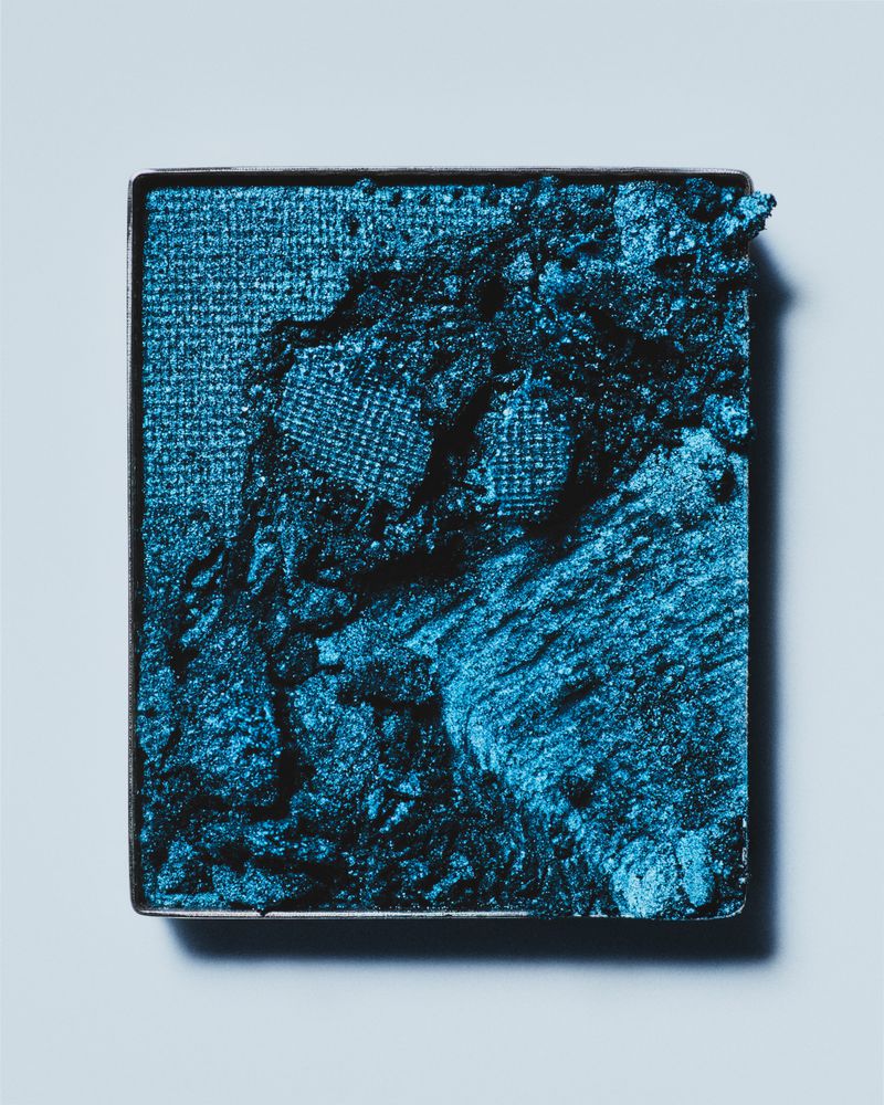 Close-up of a square container holding vibrant blue eyeshadow, featuring a textured surface and a slight shimmer on a light blue background.