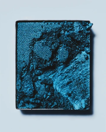 Close-up of a square container holding vibrant blue eyeshadow, featuring a textured surface and a slight shimmer on a light blue background.