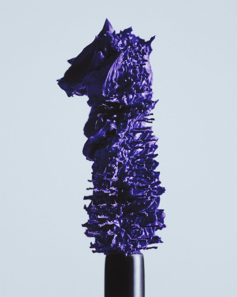 Close-up studio shot of a mascara wand covered in purple mascara against a neutral background. Beauty product photography.