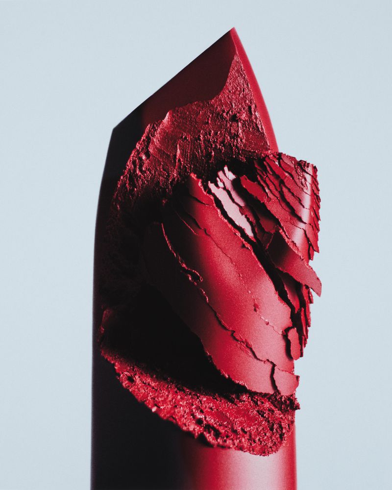 Close-up studio shot of a red lipstick with a broken tip against a gradient blue background, emphasizing textures and makeup product details.