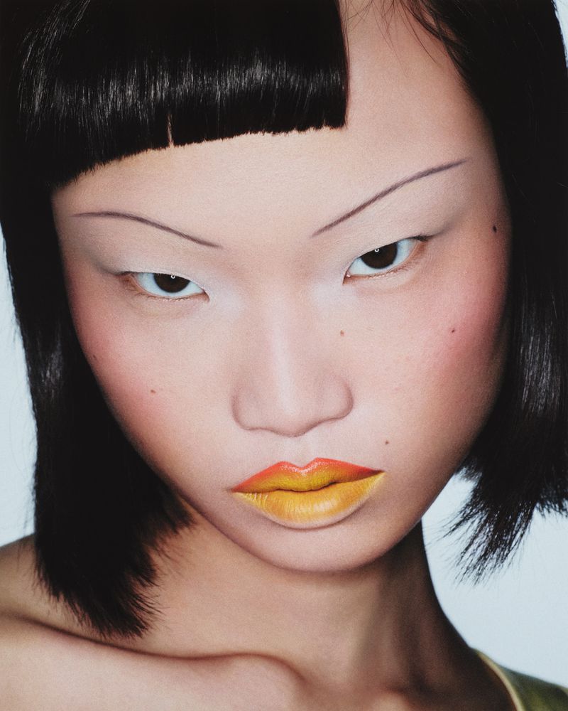 Close-up portrait of an Asian woman with black bob haircut, dramatic makeup, and yellow ombre lips in a studio environment.