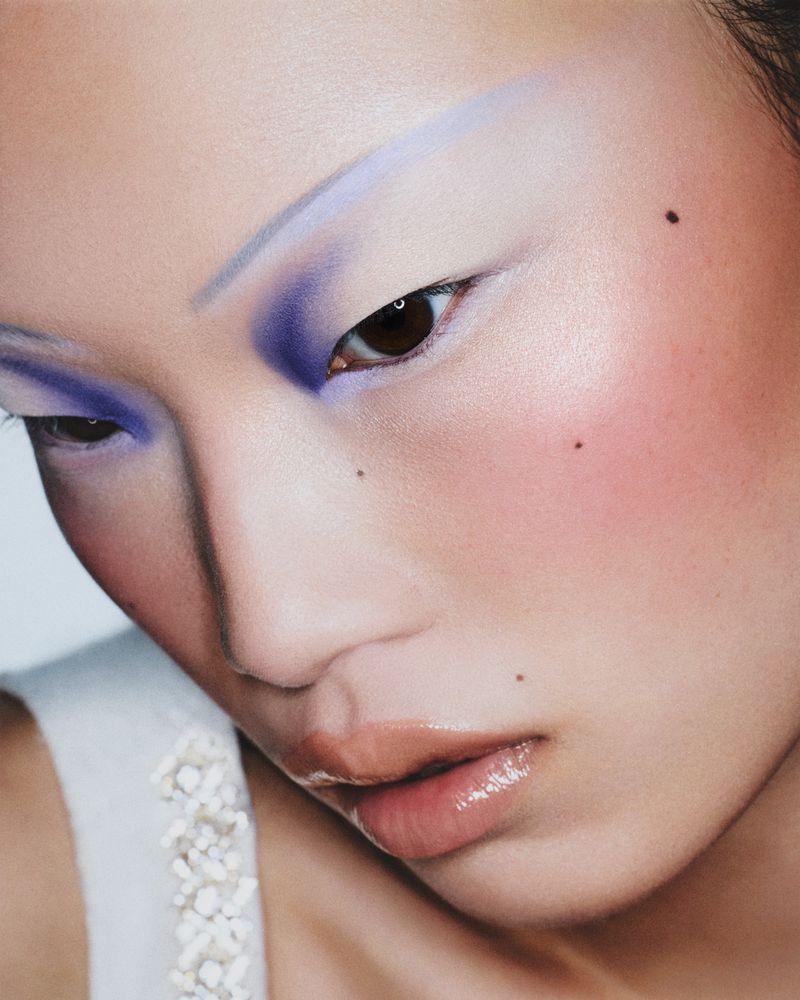 Close-up portrait of a woman with dramatic makeup featuring blue eyeshadow and glossy lips against a soft, neutral background.