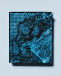 Close-up of a shattered metallic blue eyeshadow pan with textured pigment against a light background