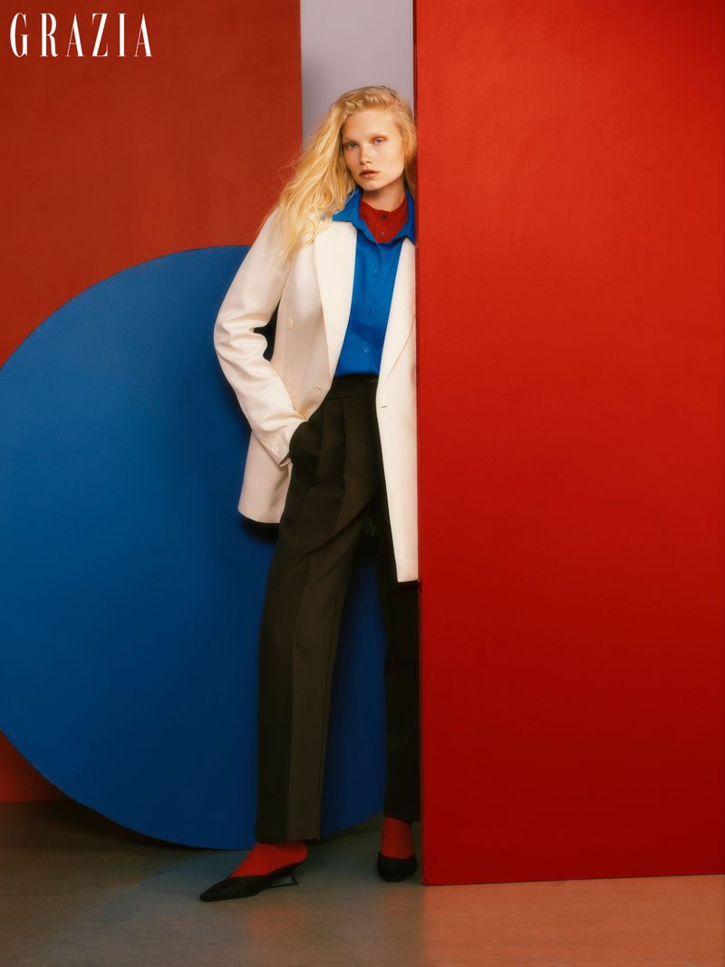 Fashion editorial shot of a blonde woman in a white blazer, blue shirt, and black trousers, posing against red and blue graphic panels.