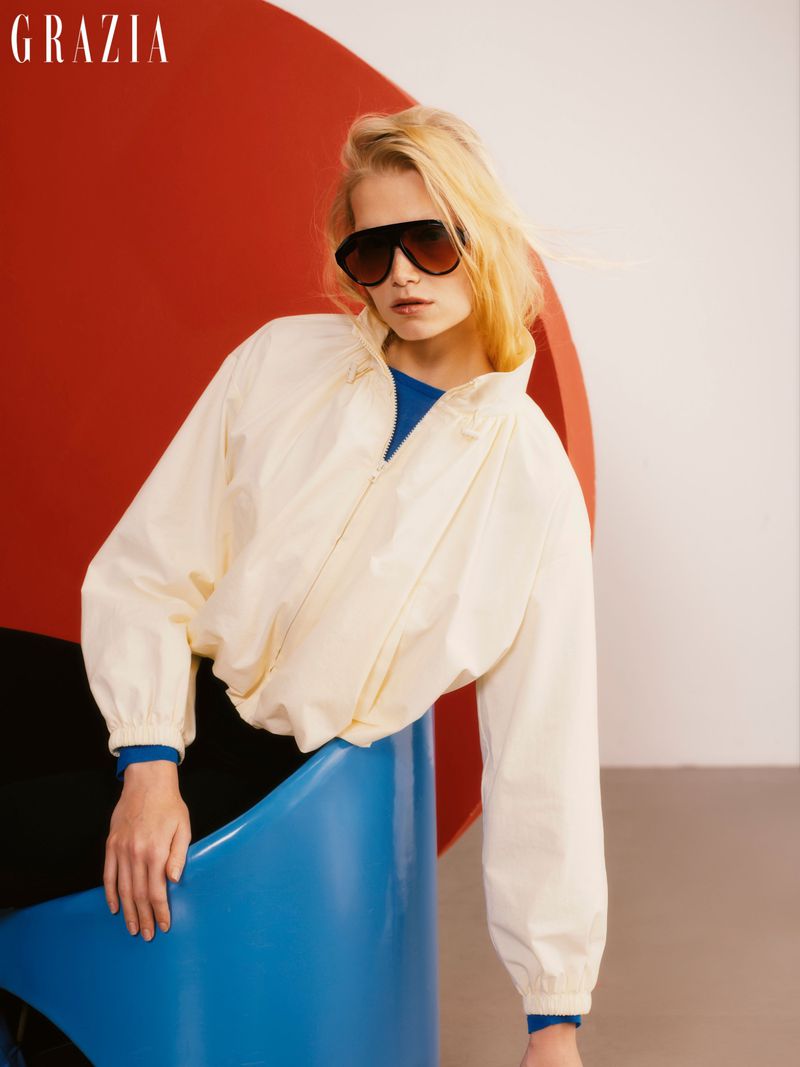 Fashion editorial shot of a fair-skinned blonde woman in an off-white jacket, sunglasses, and black pants. The setting has red, white, and blue.