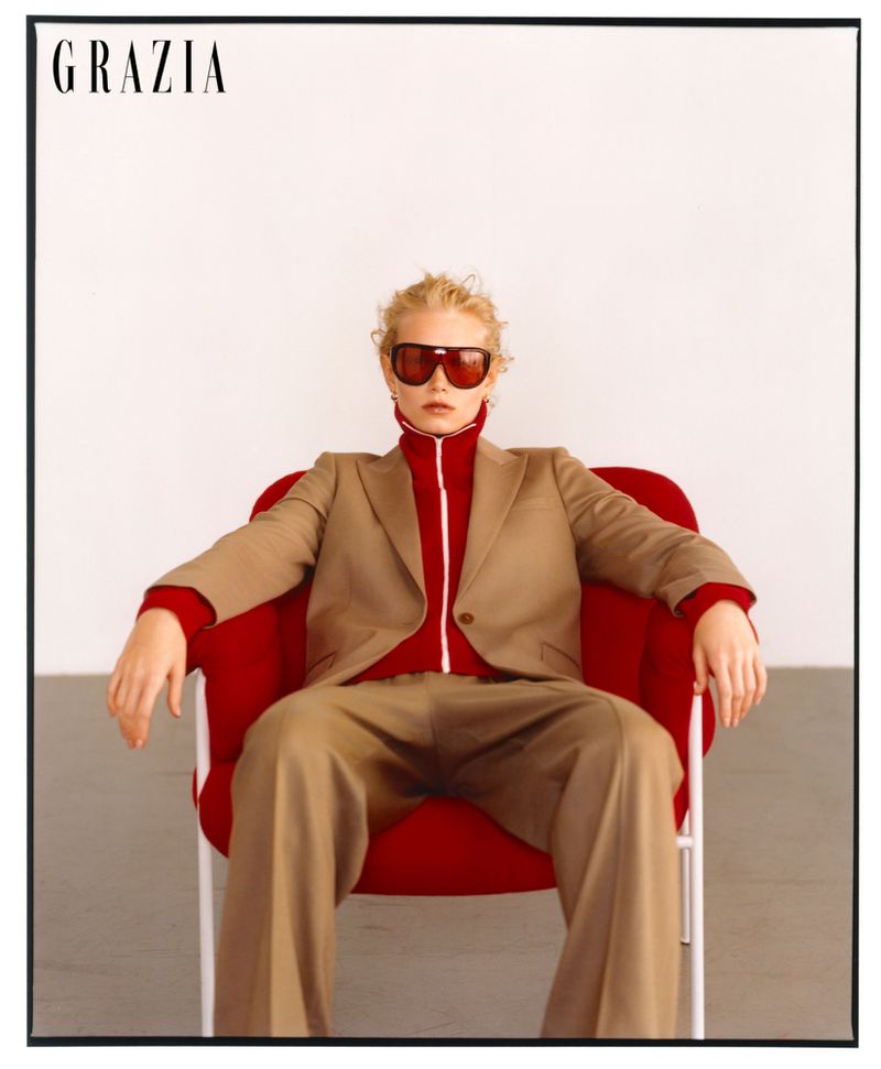 Fashion shot of a young adult woman in a beige suit and red turtleneck, seated in a red chair against a plain white background.