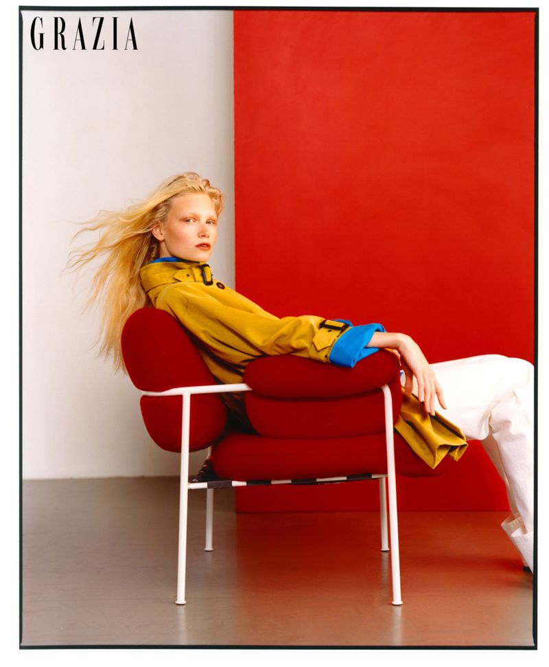 Fashion editorial shot of a blond woman in a yellow jacket and white pants lounging in a red chair against a split white and red backdrop.