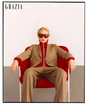 Model wearing a tan suit and sunglasses seated in a red armchair against a plain white background