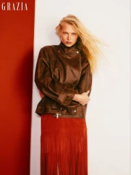 Model with blonde hair wearing a brown leather jacket and red fringed skirt against a white and red background.